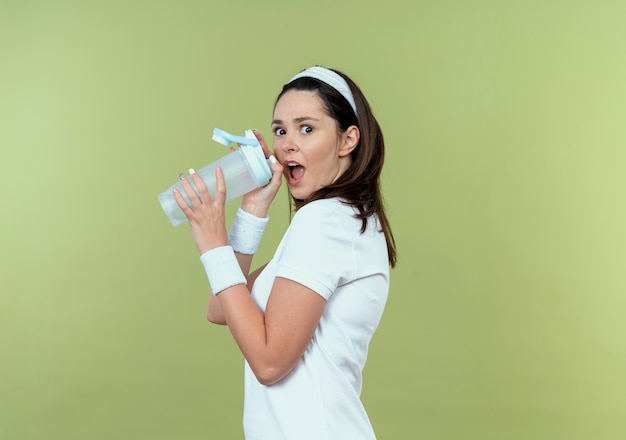 Junge Fitnessfrau im Stirnband, die Flasche Wasser überrascht über Stehwand hält