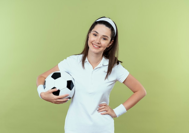 Junge Fitnessfrau im Stirnband, die Fußball mit Arm an der Hüfte hält, die glücklich und positiv über Lichtwand lächelnd lächelt