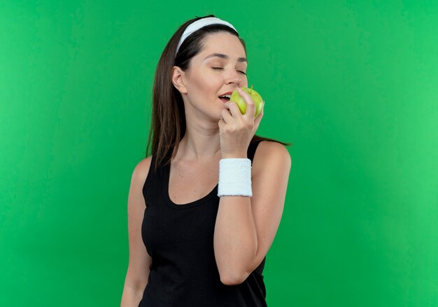 junge Fitnessfrau im Stirnband, die grünen Apfel hält, der es glücklich und positiv beißend über grüner Wand beißt
