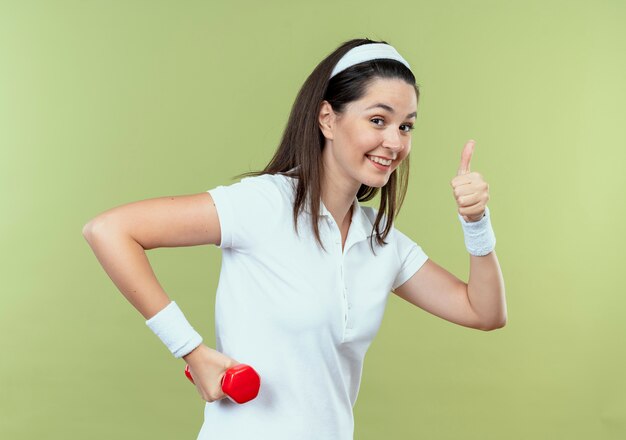 Junge Fitnessfrau im Stirnband, die mit Hantel arbeitet, die Kamera lächelnd zeigt, zeigt Daumen hoch stehend über hellem Hintergrund