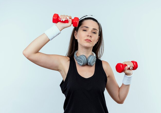 Junge Fitnessfrau im Stirnband, die mit Hanteln arbeitet, die müde stehen über weißem Hintergrund stehen