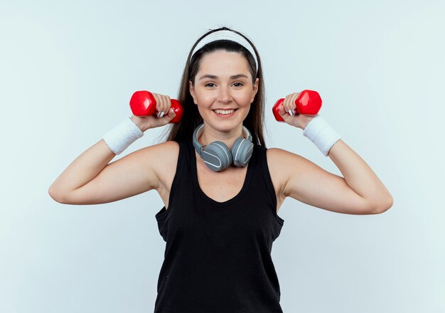 Junge Fitnessfrau im Stirnband, die mit Hanteln arbeitet, die sicher lächelnd über weißem Hintergrund stehend schauen