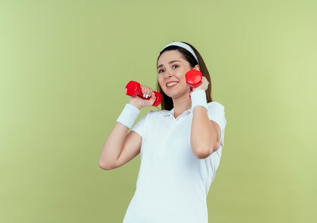 Junge Fitnessfrau im Stirnband, die mit Hanteln arbeitet, die verwirrt stehen über hellem Hintergrund stehen