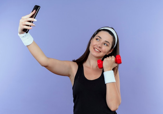 junge Fitnessfrau im Stirnband, die selfie unter Verwendung der Smartphone-Hantel hält, die mit dem glücklichen Gesicht lächelnd über der blauen Wand hält
