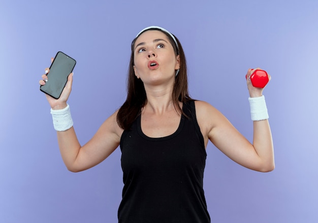 junge Fitnessfrau im Stirnband, die Smartphone und Hantel hält, die verwirrt stehen über blauer Wand stehen