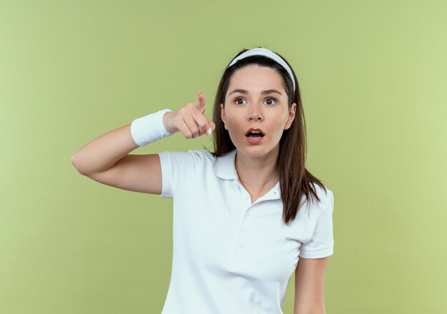 Junge Fitnessfrau im Stirnband, die überrascht mit dem Finger auf etwas steht, das über hellem Hintergrund steht