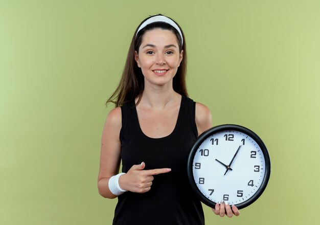 junge Fitnessfrau im Stirnband, die Wanduhr hält, zeigt mit dem Finger auf sie lächelnd stehend über Lichtwand