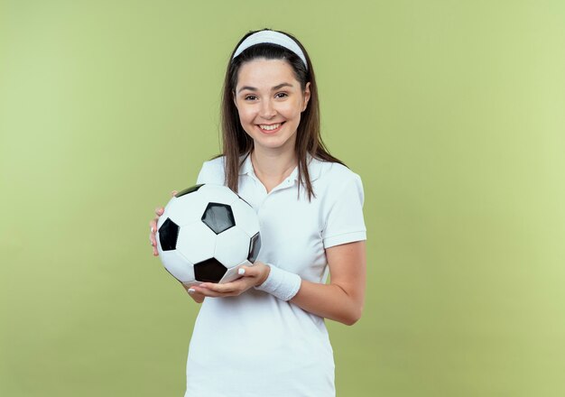 junge Fitnessfrau im Stirnband hält Fußball lächelnd glücklich und positiv stehend über Lichtwand