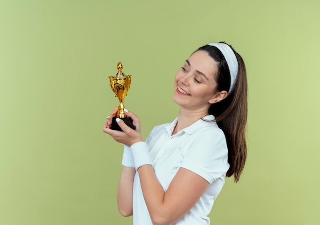 Kostenloses Foto junge fitnessfrau im stirnband hält ihre trophäe und schaut sie lächelnd mit glücklichem gesicht an, das über lichtwand steht