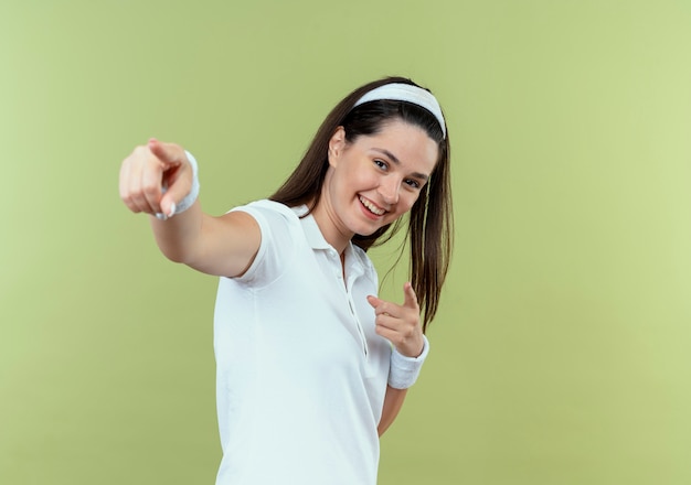 Junge Fitnessfrau im Stirnband lächelnd, das mit dem Finger zur Kamera steht, die über hellem Hintergrund steht