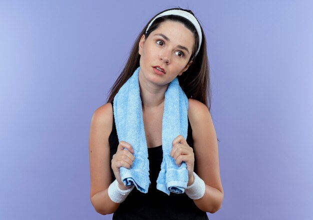 Junge Fitnessfrau im Stirnband mit Handtuch um den Hals, der müde und gelangweilt über der blauen Wand beiseite schaut