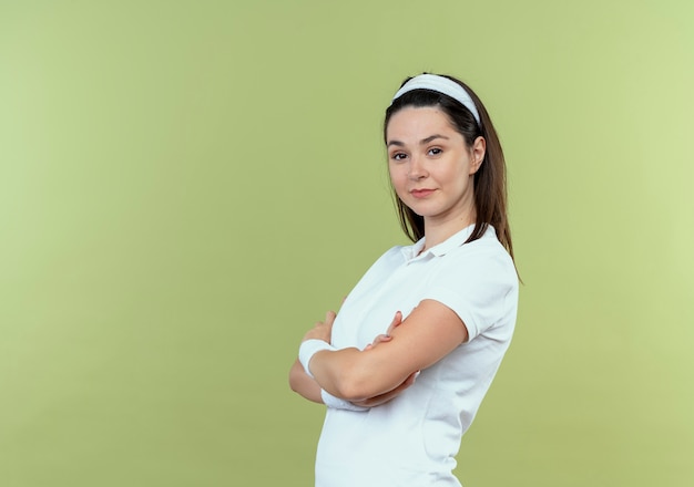 junge Fitnessfrau im Stirnband mit selbstbewusstem Ausdruck mit verschränkten Armen über heller Wand stehend