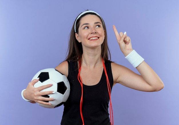 Junge Fitnessfrau im Stirnband mit Springseil um den Hals, der Fußball zeigt, der oben mit dem Finger zeigt, der oben lächelnd neue Idee hat, die über blaue Wand steht