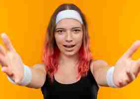 Kostenloses Foto junge fitnessfrau in der sportbekleidung, die begrüßungsgeste mit den händen macht, die über orange wand stehen