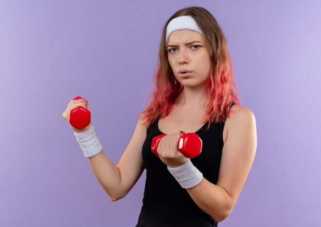 Junge Fitnessfrau in der Sportbekleidung, die Übungen unter Verwendung der Hanteln mit dem ernsten Gesicht belastet macht, das über lila Wand steht