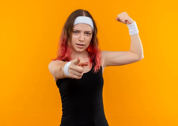 Junge Fitnessfrau in der Sportbekleidung, die Faust schaut, die sicher zeigt, mit Zeigefinger auf Kamera stehend über orange Wand zeigt