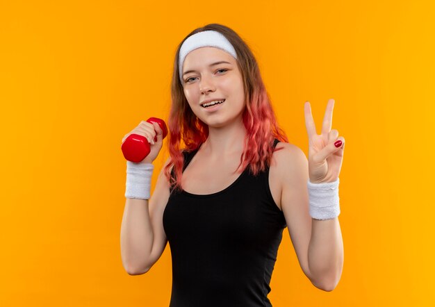 Junge Fitnessfrau in der Sportbekleidung, die Hantel lächelnd fröhlich zeigt Siegzeichen steht über orange Wand