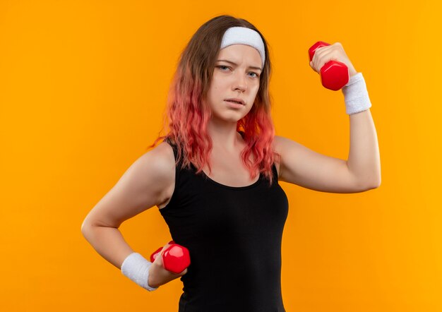 Junge Fitnessfrau in der Sportbekleidung, die Hanteln hält, die zuversichtlich zeigen, dass Stärke über orange Wand steht