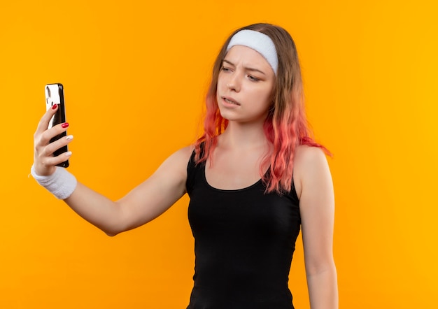 Junge Fitnessfrau in der Sportbekleidung, die selfie unter Verwendung des Smartphones mit ernstem Gesicht steht über orange Wand nimmt