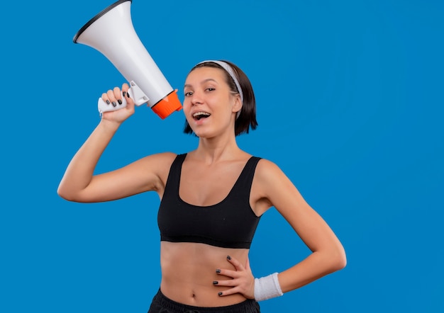 Junge Fitnessfrau in der Sportbekleidung, die zum Megaphon schreit, das glücklich und zuversichtlich steht, über blauer Wand stehend
