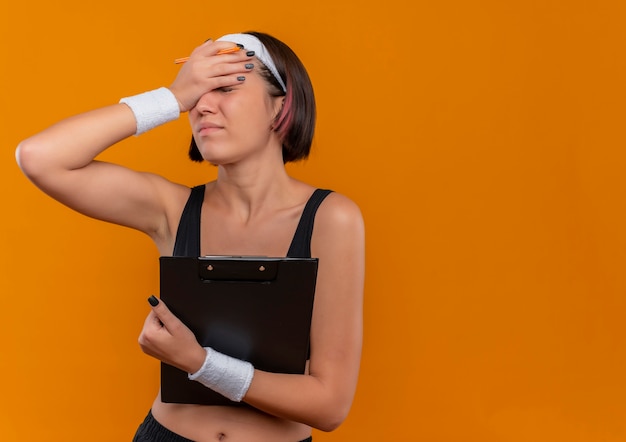 Kostenloses Foto junge fitnessfrau in der sportbekleidung mit dem stirnband, das klemmbrett hält, das müde und gelangweilte bedeckende augen mit der hand steht über orange wand schaut