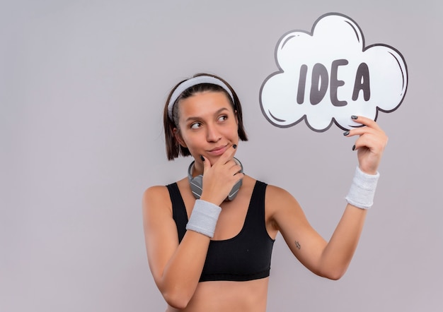 Junge Fitnessfrau in der Sportbekleidung mit dem Stirnband, das Sprachblasenzeichen mit Wortidee hält, die beiseite mit nachdenklichem Ausdruck steht, der über weißer Wand steht