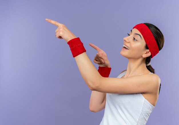 Junge Fitnessfrau in der Sportbekleidung mit dem Stirnband, das überrascht und erstaunt beiseite schaut und mit dem Finger zur Seite zeigt, die über lila Wand steht
