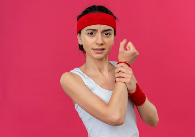 Junge Fitnessfrau in der Sportbekleidung mit dem Stirnband, das unwohl schaut, das ihr Handgelenk berührt, das Schmerzen hat, die über rosa Wand stehen