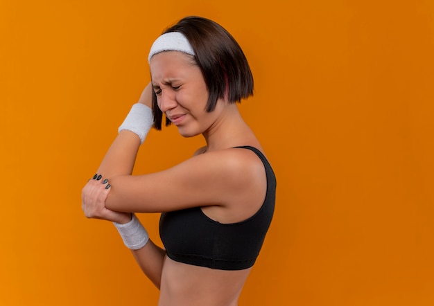 Junge Fitnessfrau in der Sportbekleidung mit dem Stirnband, das unwohl schaut, das ihren Ellbogen berührt, der Schmerz fühlt, der über orange Wand steht