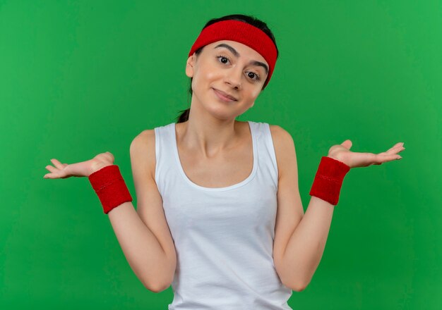 Junge Fitnessfrau in der Sportbekleidung mit dem Stirnband, das verwirrte und unsichere achselzuckende Schultern sieht, die keine Antwort über grüner Wand stehen