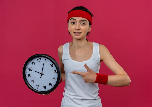 Junge Fitnessfrau in der Sportbekleidung mit dem Stirnband, das Wanduhr hält, zeigt mit dem Finger, der verwirrt steht über rosa Wand