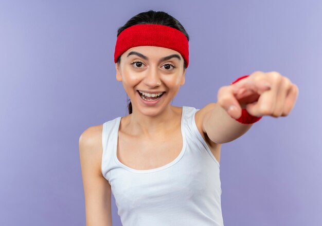 Junge Fitnessfrau in der Sportbekleidung mit dem Stirnband, der fröhlich mit dem Zeigefinger zur Kamera zeigt, die über lila Wand steht