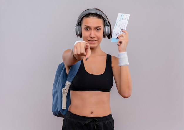 Junge Fitnessfrau in der Sportbekleidung mit Kopfhörern auf Kopf mit Rucksack, der Flugschein hält, zeigt mit dem Finger auf Kamera lächelnd über weiße Wand stehend