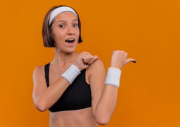 Junge Fitnessfrau in der Sportbekleidung mit positivem und glücklichem Stirnband mit Daumen, die über orange Wand stehen