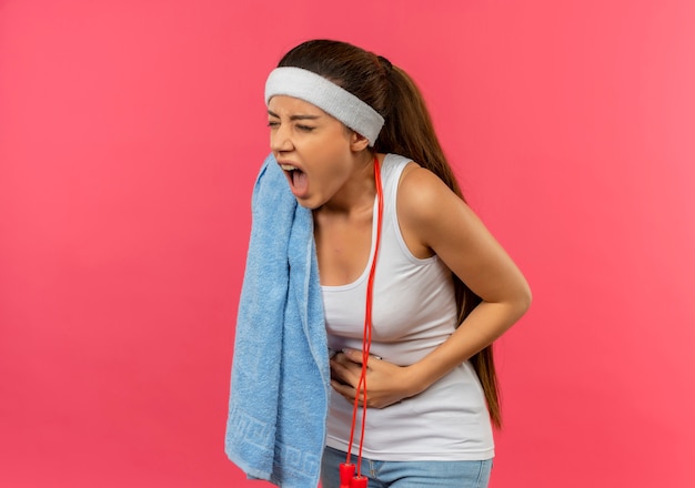 Junge Fitnessfrau in der Sportbekleidung mit Stirnband und Handtuch auf ihrer Schulter, die ihren Bauch berührt, der unwohl fühlt, der Schmerz steht über rosa Wand