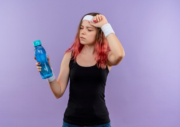 Junge Fitnessfrau in der Sportkleidung, die Flasche Wasser hält, die müde steht über lila Wand steht