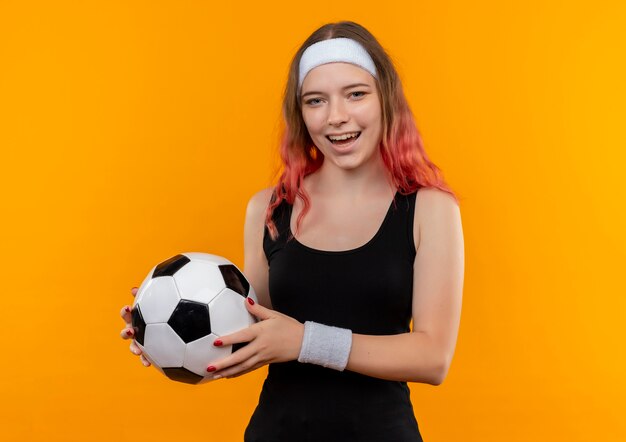Junge Fitnessfrau in der Sportkleidung, die Fußball mit glücklichem Gesicht hält, das fröhlich über orange Wand steht