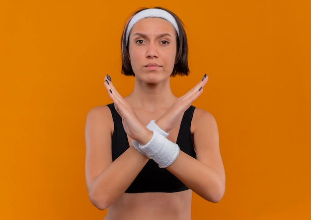 Kostenloses Foto junge fitnessfrau in der sportkleidung mit dem stirnband, das hände kreuzt, die verteidigungsgeste stellen, die über orange wand steht