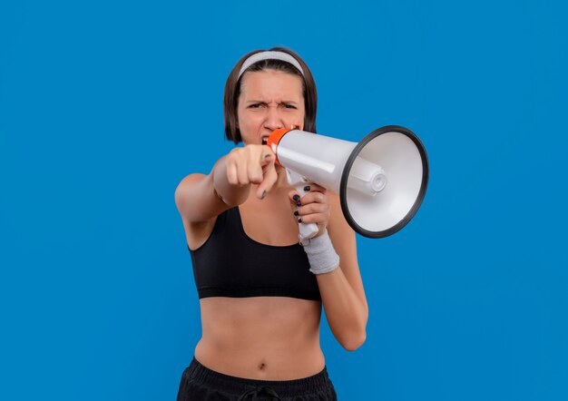 Junge Fitnessfrau in Sportbekleidung, die zum Megaphon mit aggressivem Ausdruck schreit, der mit Zeigefinger zur Kamera zeigt, die über blauer Wand steht