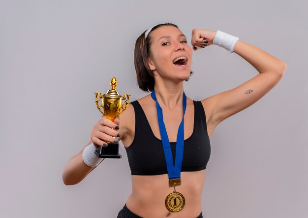 Junge Fitnessfrau in Sportbekleidung mit Goldmedaille um den Hals hält ihre Trophäe, die die Faust hebt, glücklich und aufgeregt und freut sich über ihren Erfolg, der über weißer Wand steht