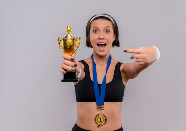 Junge Fitnessfrau in Sportbekleidung mit Goldmedaille um den Hals hält ihre Trophäe, die mit dem Finger darauf zeigt, glücklich und aufgeregt und freut sich über ihren Erfolg, der über weißer Wand steht