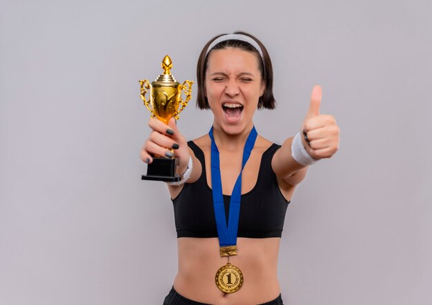 Junge Fitnessfrau in Sportbekleidung mit Goldmedaille um den Hals hält ihre Trophäe glücklich und aufgeregt und freut sich über ihren Erfolg, Daumen hoch stehend über weißer Wand