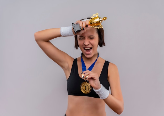 Kostenloses Foto junge fitnessfrau in sportbekleidung mit goldmedaille um den hals hält ihre trophäe glücklich und aufgeregt und freut sich über ihren erfolg, der über weißer wand steht