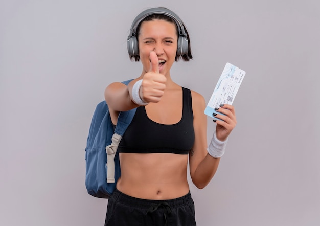 Junge fitnessfrau in sportbekleidung mit kopfhörern auf kopf mit rucksack, der flugschein lächelnd zeigt daumen hoch stehend über weißer wand