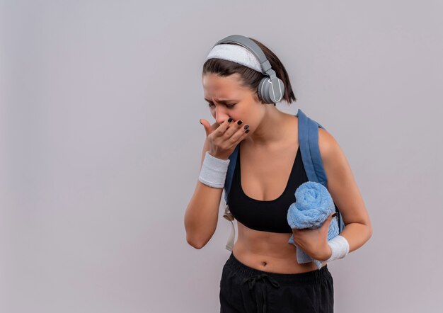 Junge Fitnessfrau in Sportbekleidung mit Rucksack und Kopfhörern auf Kopf, der Handtuch hält, das unwohl hustend steht über weißer Wand steht