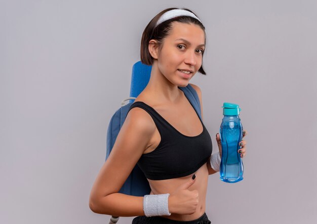 Junge Fitnessfrau in Sportbekleidung mit Rucksack und Yogamatte, die Flasche Wasser lächelnd zeigt Daumen hoch stehend über weißer Wand