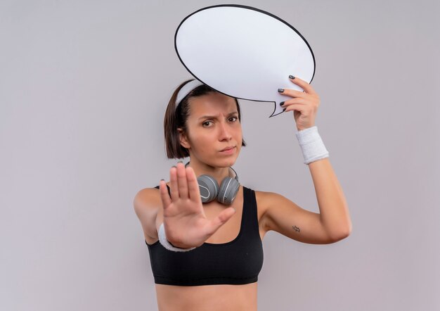 Junge Fitnessfrau in Sportbekleidung mit Stirnband, das leeres Sprachblasenzeichen hält Stoppschild mit Hand mit ernstem Gesicht, das über weißer Wand steht