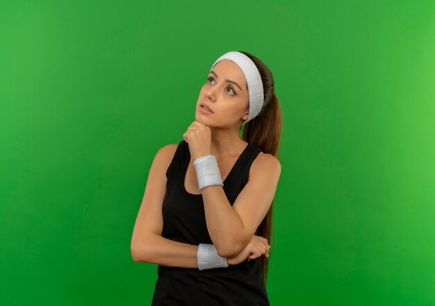 Junge Fitnessfrau in Sportbekleidung mit Stirnband, das mit Hand auf Kinn mit nachdenklichem Ausdruck beiseite schaut, der über grüner Wand steht