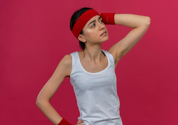 Junge Fitnessfrau in Sportbekleidung mit Stirnband, das verwirrt und sehr ängstliches Denken beiseite schaut und Zweifel hat, die über rosa Wand stehen
