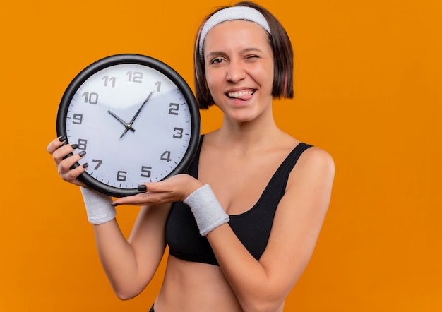 Junge Fitnessfrau in Sportbekleidung mit Stirnband, das Wanduhr glücklich positiv herausstehende Zunge steht über orange Wand hält